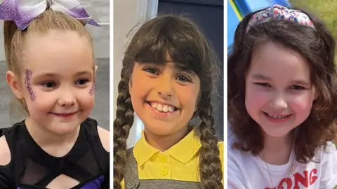  Elsie Dot Stancombe with a purple and white bow in her tied-back hair and glitter around her eyes; Alice Da Silva Aguiar with brown braids, a yellow top and a grey dress; and Bebe King with a multi-coloured headband and a white t-shirt.