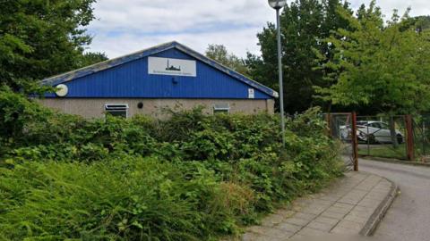 A single-storey building with blue cladding stands behind dense greenery and shrubs beside a narrow road. A sign on the front of the building reads “Borehamwood Islamic Society”. A lamppost stands in the foreground, while a metal gate to the right leads to a parking area where a white car is visible. Trees surround the site under a cloudy sky.
