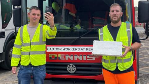 Two workers for Barnsley's waste collection service, stood in front of a bin lorry, wearing high visibility jackets. One holds aloft a mobile phone. The other holds the technology used to track mobile signal coverage as the lorries traverse the area - which looks like a white box.