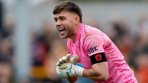 Goalkeeper Jack Sims, pictured in the pink goalkeeping strip at Sutton United, shouts and claps hands.