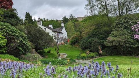 Rydal Mount, a large white country cottage with a grey roof and several chimney stacks, stands in the background of the image, surrounded by trees and grass. A number of bluebells can be seen in the foreground.