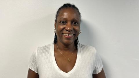 A woman with black hair and wearing a white T-shirt and a gold necklace smiling at the camera against a plain white background.