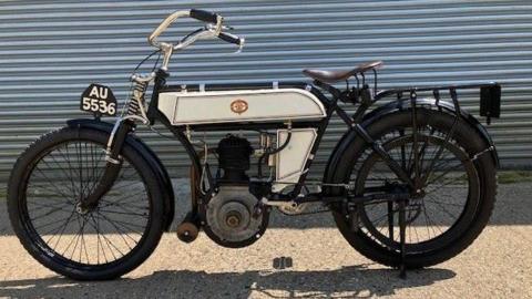 A black and white 1904 Riley motorcycle, parked in front of a corrugated silver shutter.