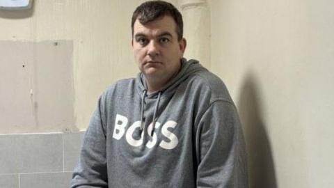 Andrew Gibney has short black hair and is wearing a grey hoodie with BOSS white letters logo.