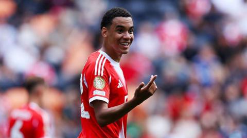 Jamal Musiala of Bayern Munich, in red kit, smiling and making a signal with his right hand