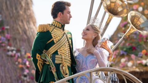 Jonathan Bailey in character as Fiyero and Ariana Grande as Glinda, as they appear in Wicked: For Good. They are looking into each other's eyes, stood on a stage in front of a floral background.