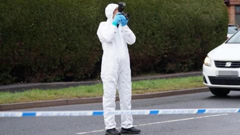 A person in a white boiler suit stood inside a police tape cordon. The person is wearing blue gloves and holding a camera up to their face.