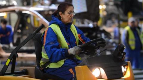 A woman driving a vehicle inside  a factory, wearing a hi-vis vest on top of work overalls. 