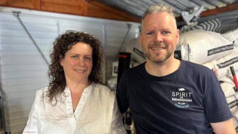 A woman with brown curly hair is looking at the camera smiling. She is wearing a white v-neck shirt. Next to her is a man with short grey hair who is also smiling at the camera. He is wearing a blue t-shirt with "Spirit of Birmingham" on it. Behind them is a white garage door and white bags of barley.