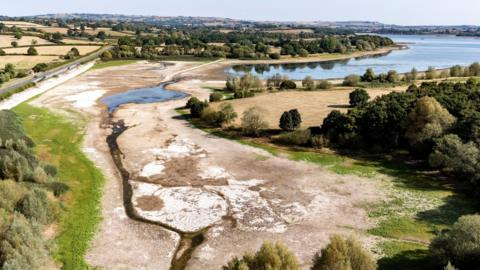 An aerial image showing the Chew Valley Lake at half-full capacity during August. The section of the reservoir nearest to the camera shows a dry brown lake bed with a small tributary running through it. In the distance, as the basin gets deeper, there is more water towards the right hand side of the image. The entire lake is surrounded by dry brown fields and thick green trees. 