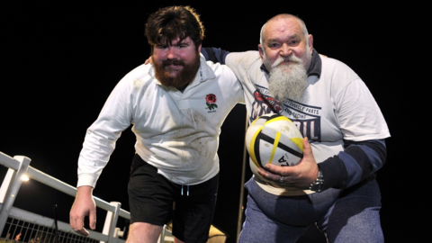 Joseph and Graeme on a flood-lit rugby pitch at night in white kit. Graeme has his arm around his son and is holding a ball. Both are slightly crouched down as if in action