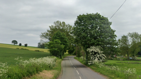 A general view of the B966 at Cairnton of Balbegno. The road cuts through the middle of the image and is grey with white markings on it. There is grass and fields on either side and in the middle of the image, trees going overhead. They sky is grey.