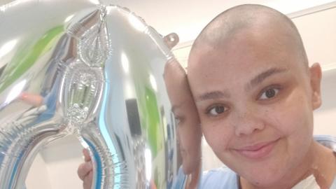 Lakita has lost her hair through chemo and stands smiling in a hospital room, holding a silver balloon shaped like a 0. Her reflection is partly visible in the balloon.