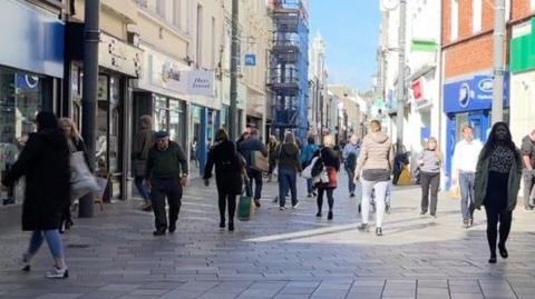 People walking through Strand street in Douglas at lunchtime. They are a mix of ages and genders, with some carrying bags. The street is paved and there is shop signage on either side.