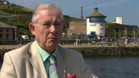 Graham Roberts stands on a seafront in Whitehaven, Cumbria. He is elderly and wears a beige suits and a mint-coloured shirt with a matching tie. 