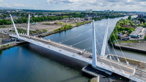 A white bridge across a river with empty industrial land on one side and factories on the other bank