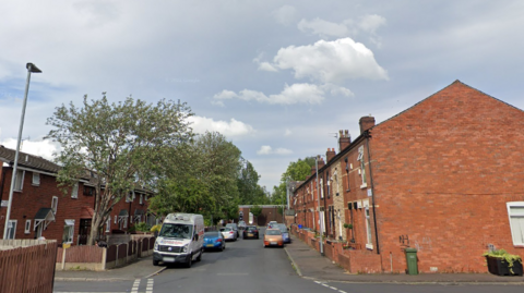 A screenshot from Google streetview of a residential street with red brick houses and cars parked on either side of the road. 