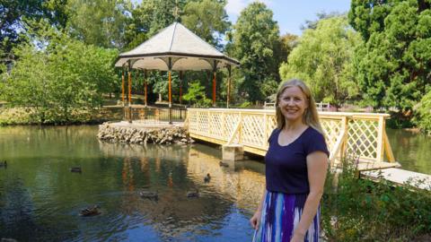 Councillor Karen Lewing at the Gheluvelt Park bandstand. The bandstand is on a rocky platform in the middle of a lake with a wooden bridge linking it to the shoreline. Lewis is wearing a navy blue top with a blue patterned skirt.
