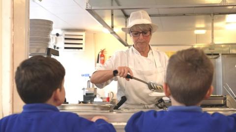 Dinner lady serving two young pupils in a canteen