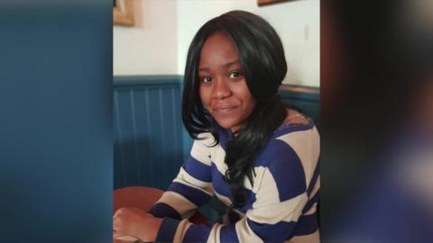 Chanel Thompson sits at a table in a café, wearing a blue and white striped top, looking at the camera.