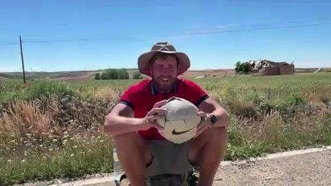 James sits on his bag amid high temperatures as he is interviewed while holding his football. He has a large sunhat on with a red T-shirt and shorts. He is unshaven. Grassland can be seen behind him and a blue, cloudless sky.