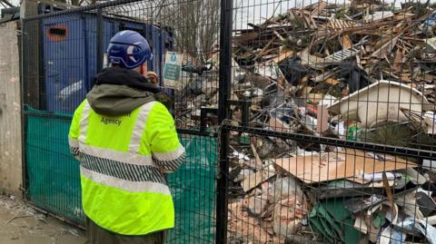 A man in a hi-vis Environment Agency Jacket and a hard hat stands in front of locked gates at the waste site. The waste is piled up and includes timber and other debris. There are trees in the background,