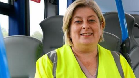 Councillor Denise Rollo wearing a hi-vis jacket and sitting on a bus. She has blonde shoulder-length hair and is smiling at the camera.