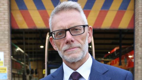 Darren Roe wearing a blue suit, a blue shirt and blue tie. He has black square-framed glasses and is looking directly at the camera. He has white hair and a white beard. 