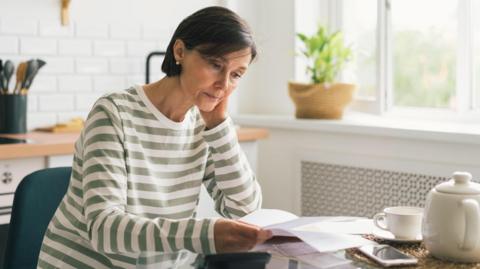 A stock image of a woman with short brown hair, wearing a green and white stripy top. She is sitting at a table holding bits of paper, which are being made to look like energy bills.