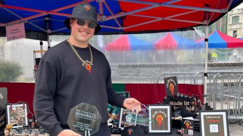 A man holding a glass trophy, stood in front of a market stall. He is wearing a black bucket hat and jumper with a red cartoon chilli on it. He has a large gold chain around his neck.