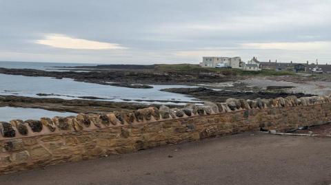 The coastal defence wall along Harbour Road in Beadnell, Northumberland. The stone wall is built near the shore. The sea can be seen behind it. A village can be seen in the distance.
