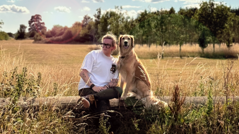 Kate is pictured wearing black-rimmed glasses and a white T-shirt. She is sitting on a log in front of a large green field bordered by trees. She is sitting with her golden retriever dog Milo who is next to her. It is a bright and sunny day.