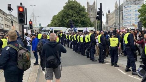 "A street protest scene with a line of police officers in high-visibility vests forming a barrier. Police vans are parked in the background, and a historic building under renovation is visible on the right, with scaffolding and construction materials present.