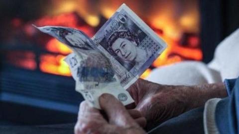 An elderly man clasps two £20 notes as he sits next to a fire.