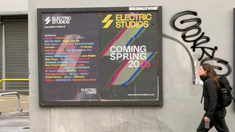 A huge billboard gig poster against a wall. A commuter in black with a large bag walks past.