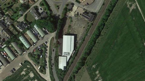 Drone image of the sit with a white warehouse with fields on side and homes on the other side.