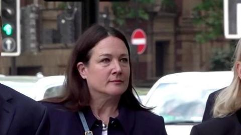 Claire Freemantle is seen with dark, shoulder-length hair and a neutral expression standing outdoors on a city street. She is wearing a dark navy jacket and a silver spherical pendant, with blurred traffic lights and road signs in the background.
