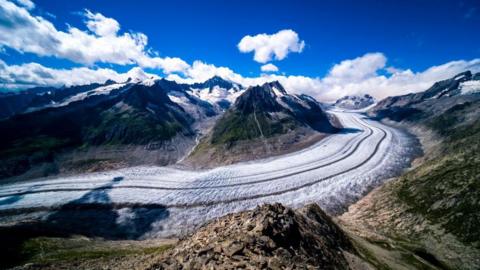 View of the Aletsch Glacier. Ice sweeps round from right to left, with mountains either side. Above is a blue sky with some clouds.