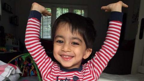 A three-year-old boy wearing red and white striped pyjamas grins at the camera. His arms are above his head and a typical family living room is visible behind him.