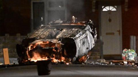 A burnt out car has been flipped over on a street with flames coming out of the front of it. There are bits of debris scattered around the car with a house seen in the background.