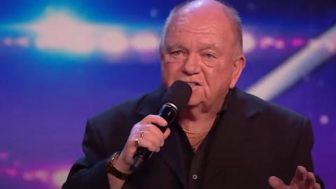 Dave Betton sings on stage during a Britain's Got Talent show. He is wearing a black jacket and shirt and holding a microphone. The background has a huge illuminated star.