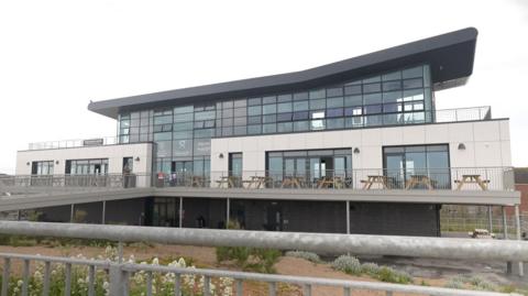 A large, modern building with white cladding, large glass windows and a sweeping black roof. The Seaview Colonnade has a terrace around it, with picnic benches behind metal railings. The building is raised off the ground, with a large raised ramp up to the entrance. There is sand and beach plants in the foreground.