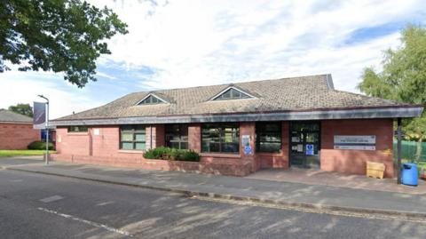 The visitor centre at HMP Hewell is a single-storey brick building with official signs and windows. It is on the corner of a road and there are trees and fields around. There is a 
