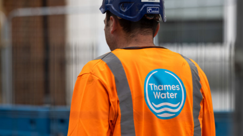 Male with back to camera wearing dark blue hardhat and high viz jacket with blue and white Thames Water logo 