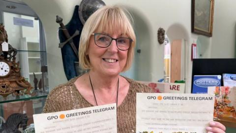 A woman smiles at the camera. She is holding two pieces of paper which have the headers "GPO Greetings Telegram". She is standing in front of multiple archways. There is a wooden clock on the left, and a sword is hung up on the wall.