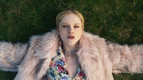 Shannon (EMMA LAIRD) in Mint, wearing a fur coat and lying on a grass lawn