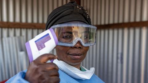 A healthworker, wearing goggles and a mask, pulled down from her mouth, holds up a thermometer