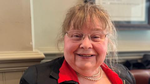 Diana is wearing pearl necklace and a black jacket with red collar