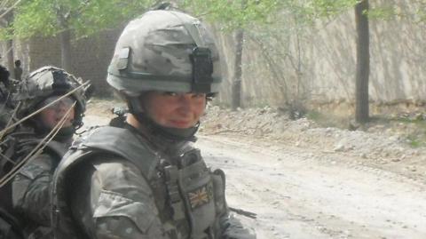 Nadine Askew wearing a camouflage military uniform and helmet. She is sitting on a dirt path beside some dry shrubs on what appears to be a military deployment. She is smiling.