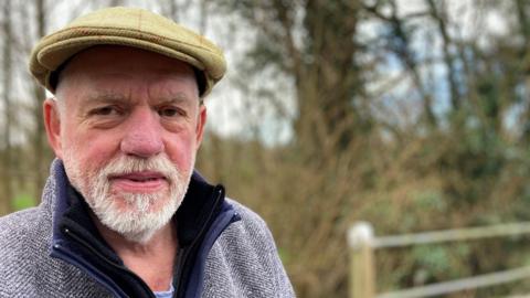 A man with grey hair and a beard in a light blue top and a light brown flat cap standing in front of a wood.